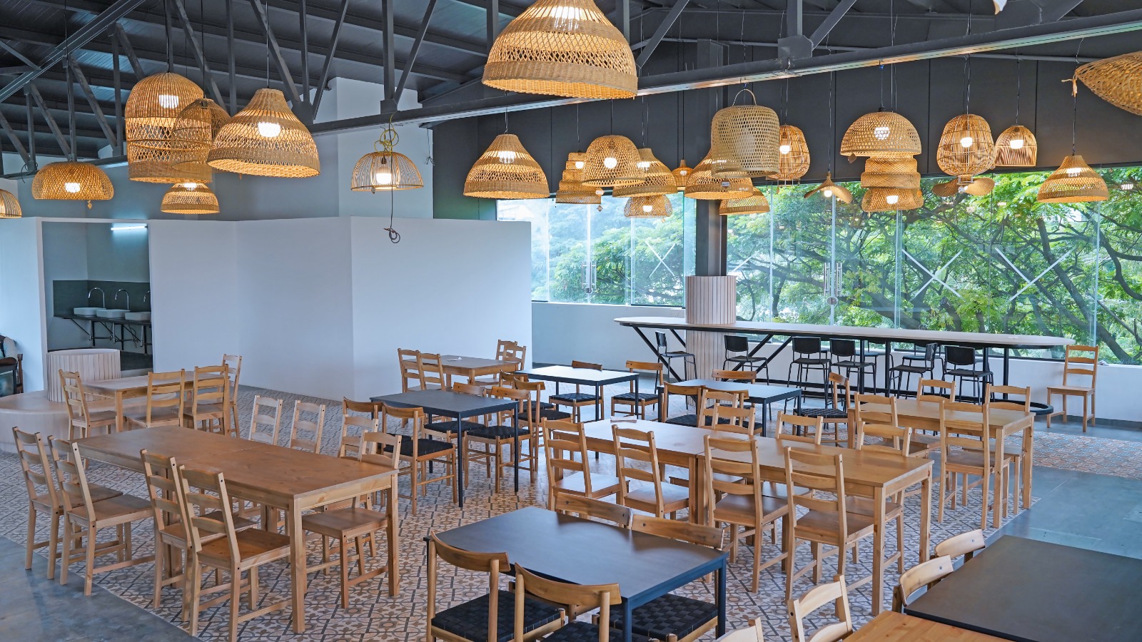 Cafeteria with long communal tables, rattan pendant lighting and full-height windows onto trees at Capstone House Bangalore